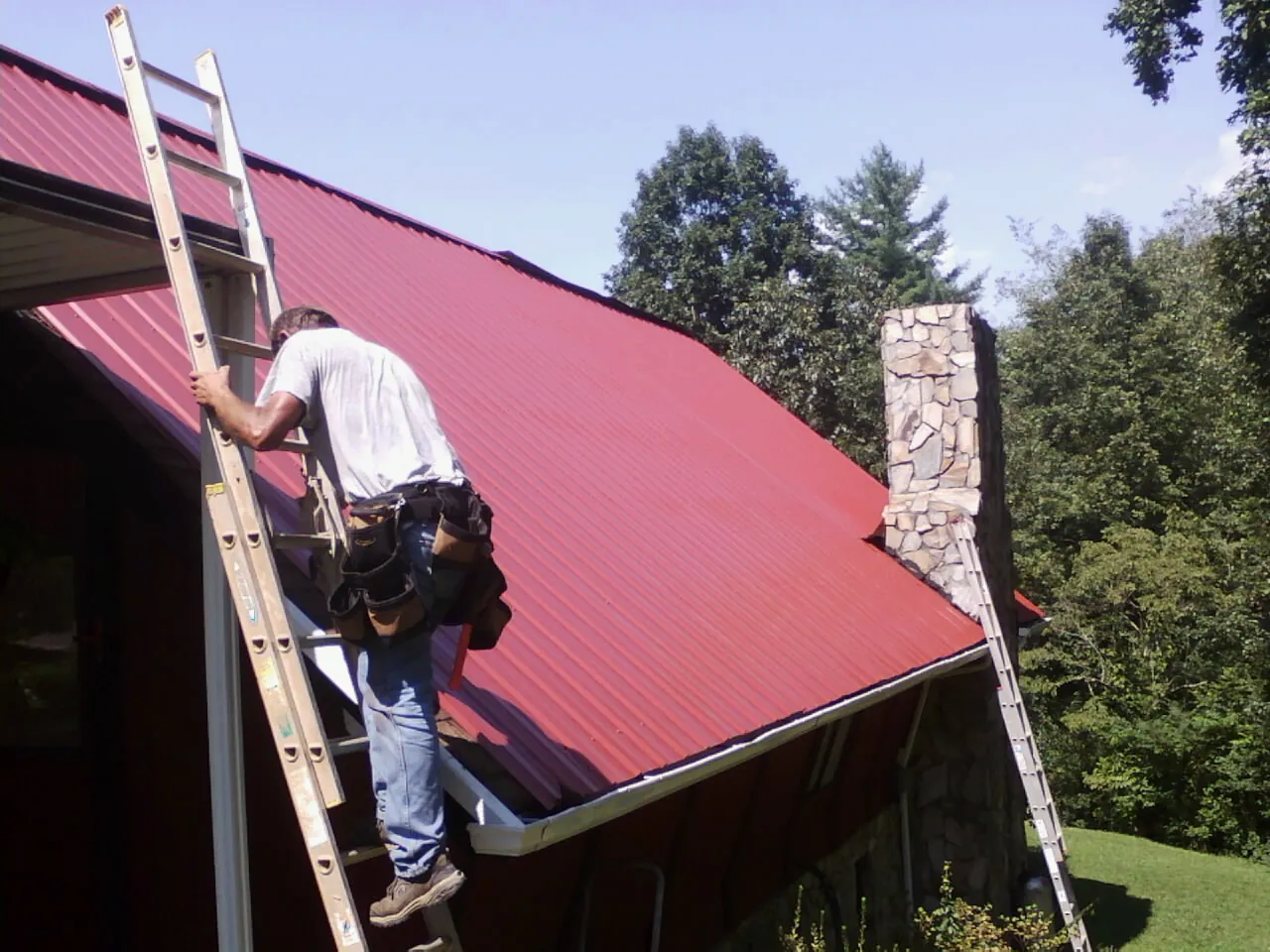Roofer on a ladder beside a red metal roof and stone chimney — Soffit & Fascia Repair by Roofing Brothers in Randall